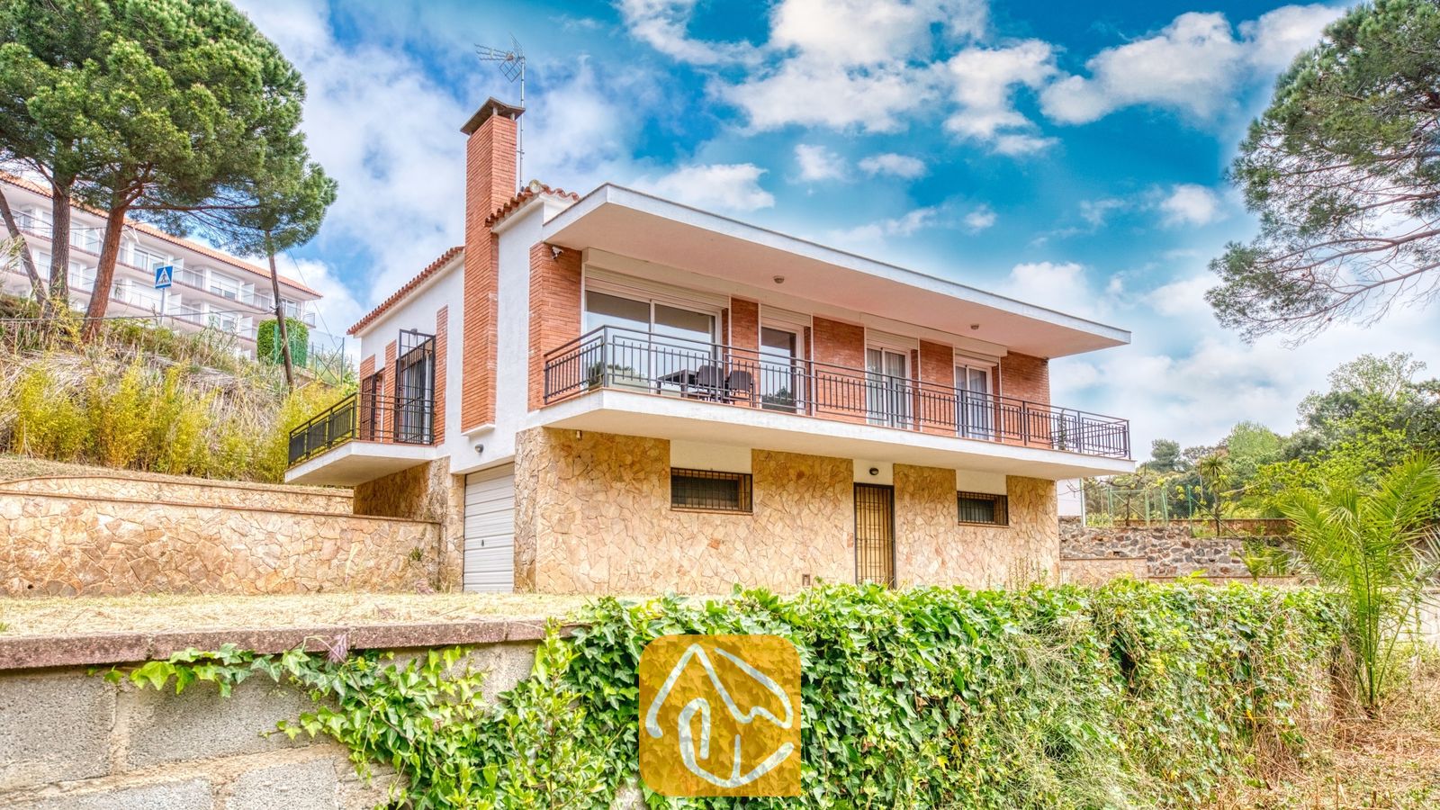 Vakantievilla's Costa Brava Spanje - Villa Lloret - Buitenaanzicht van Villa Lloret met een huis van twee verdiepingen, voorzien van een stenen benedenverdieping, een bakstenen bovenverdieping en een breed balkonterras rondom, onder een bewolkte blauwe lucht.