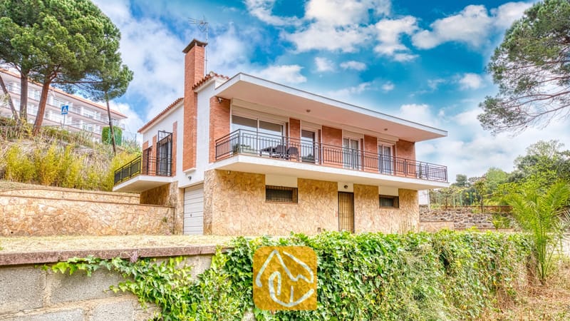 Vakantievilla's Costa Brava Spanje - Villa Lloret - Buitenaanzicht van Villa Lloret met een huis van twee verdiepingen, voorzien van een stenen benedenverdieping, een bakstenen bovenverdieping en een breed balkonterras rondom, onder een bewolkte blauwe lucht.