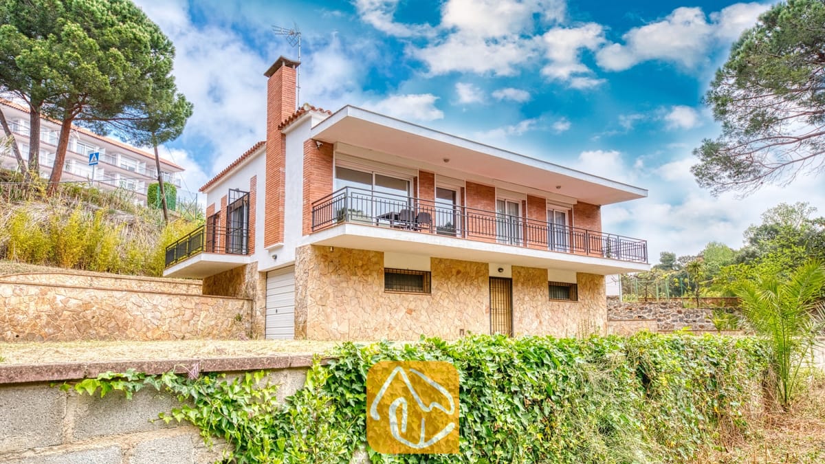 Vakantievilla's Costa Brava Spanje - Villa Lloret - Buitenaanzicht van Villa Lloret met een huis van twee verdiepingen, voorzien van een stenen benedenverdieping, een bakstenen bovenverdieping en een breed balkonterras rondom, onder een bewolkte blauwe lucht.