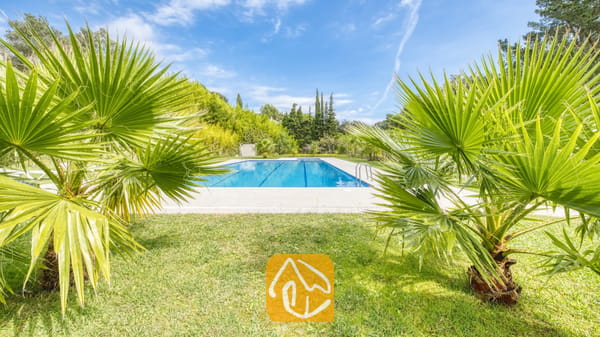 Vakantievilla's Costa Brava Spanje - Villa Lloret - Privé buitenzwembad bij Villa Lloret, omlijst door groene palmbomen, een grasveld en bomen onder een blauwe lucht.