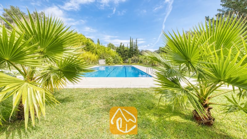 Vakantievilla's Costa Brava Spanje - Villa Lloret - Privé buitenzwembad bij Villa Lloret, omlijst door groene palmbomen, een grasveld en bomen onder een blauwe lucht.