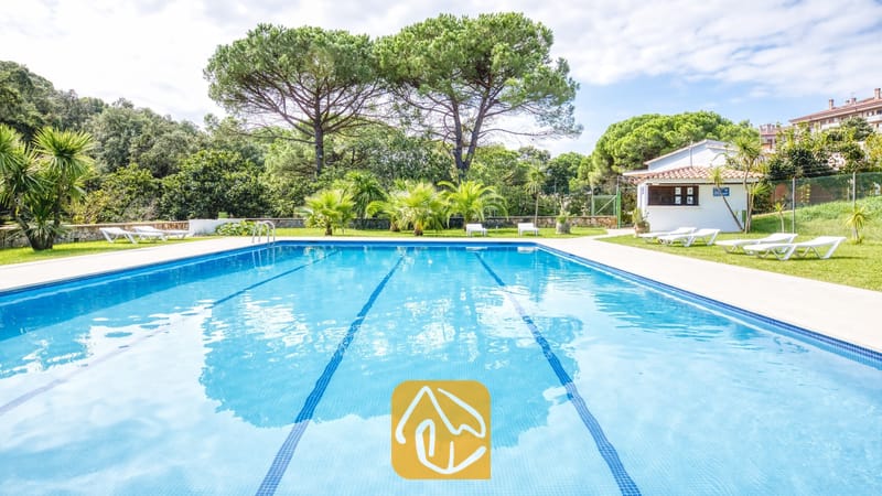 Vakantievilla's Costa Brava Spanje - Villa Lloret - Privézwembad met blauwe baanaanduidingen, omringd door een gazon, witte ligstoelen en hoge dennenbomen bij Villa Lloret.