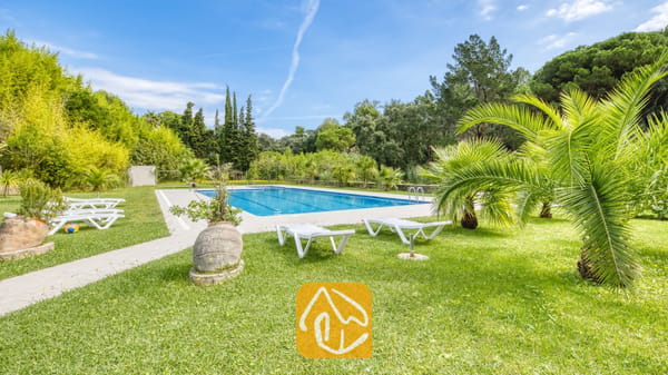 Vakantievilla's Costa Brava Spanje - Villa Lloret - Privézwembad bij Villa Lloret, omringd door groene gazons, witte plastic ligstoelen, palmbomen en dichte bosvegetatie onder een blauwe lucht.