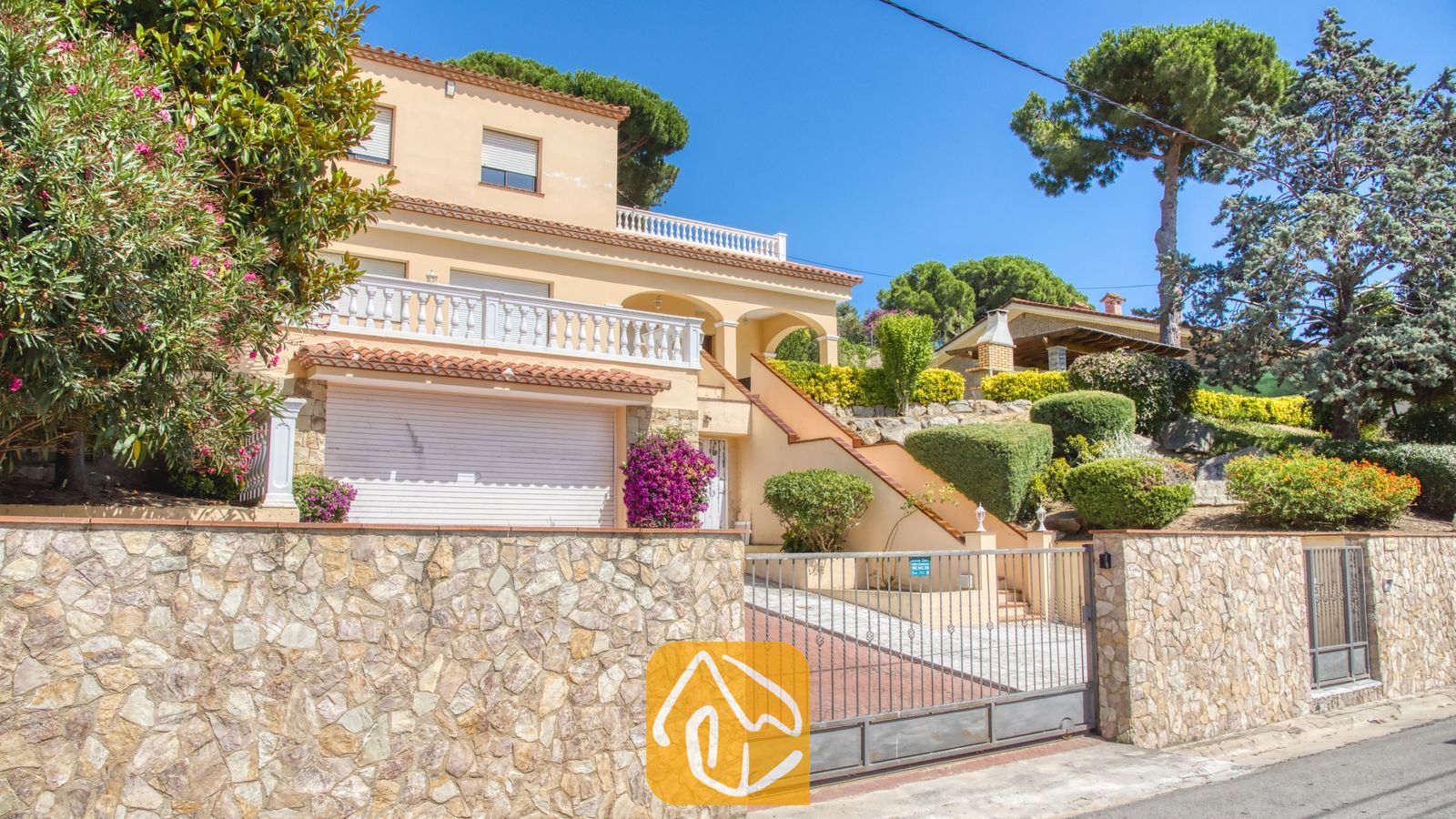 Vakantievilla's Costa Brava Spanje - Villa Paris - Buitenaanzicht van Villa Paris met de crèmekleurige gevel, witstenen balustrades, garagedeur en omringende muur van natuursteen onder een blauwe lucht.
