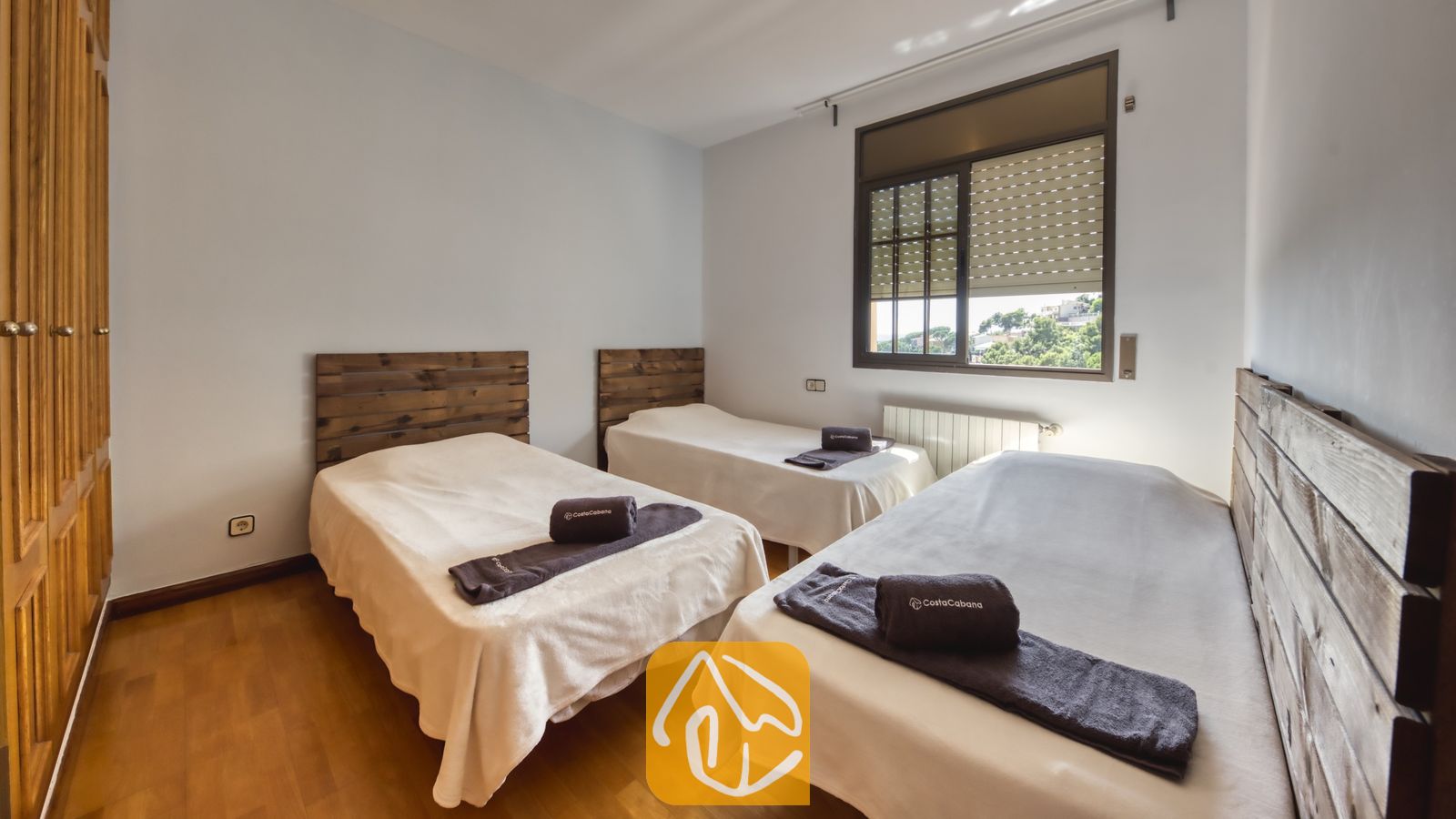 Vakantievilla's Costa Brava Spanje - Villa Paris - Slaapkamer met drie eenpersoonsbedden met rustieke houten hoofdeinden, witte beddengoed en opgerolde donkere handdoeken bovenop, tegen lichtgrijze muren en een houten vloer.