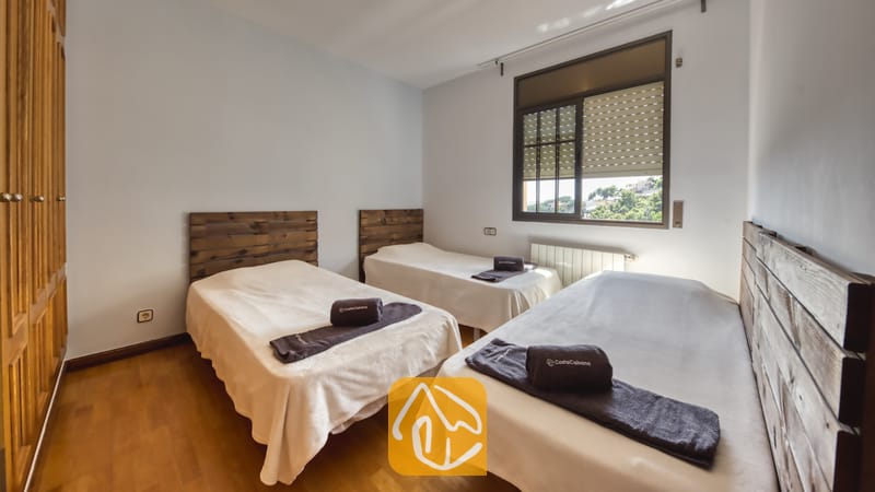 Villas de vacances Costa Brava Espagne - Villa Paris - Chambre à coucher dotée de trois lit simples avec têtes de lit en bois rustique, linge de lit blanc et serviettes sombres roulées disposées sur les lits, le tout complété par des murs gris clair et un parquet en bois.