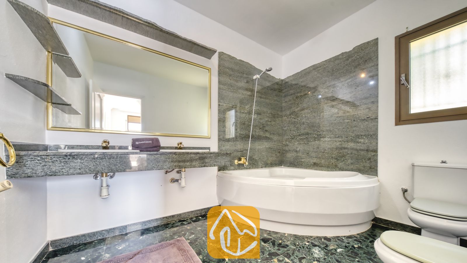 Vakantievilla's Costa Brava Spanje - Villa Paris - Badkamer voorzien van een hoekbad met wandpanelen van grijs natuursteen, een dubbele wastafel met een grote spiegel in een gouden lijst en een donkergroene marmeren vloer.