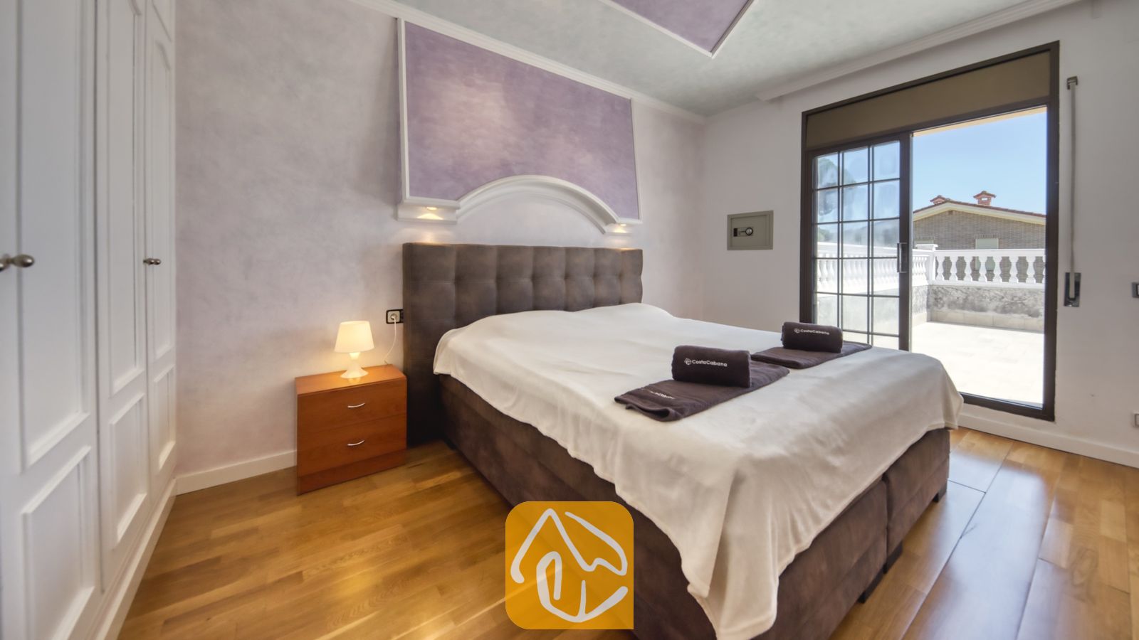 Vakantievilla's Costa Brava Spanje - Villa Paris - Slaapkamer met een groot getuft bruin hoofdbord, een houten nachtkastje met een klein lampje en een schuifpui die uitkomt op een terras.