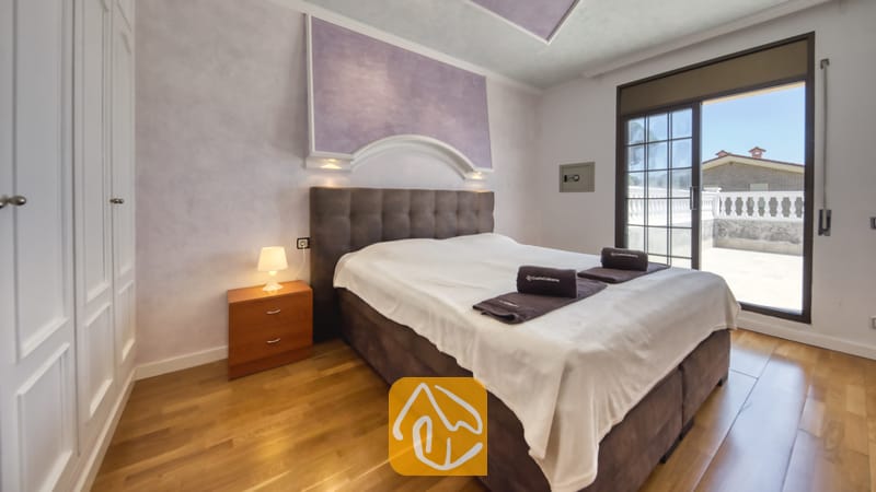 Villas de vacances Costa Brava Espagne - Villa Paris - Une chambre avec une grande tête de lit capitonnée marron, une table de chevet en bois surmontée d'une petite lampe, et une porte-fenêtre coulissant sur une terrasse.