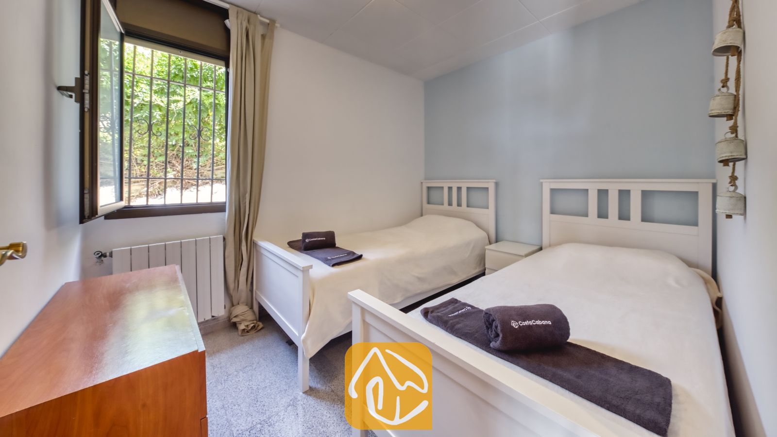 Vakantievilla's Costa Brava Spanje - Villa Paris - Slaapkamer met twee witte, houten eenpersoonsbedden, opgevouwen bruine handdoeken en een open raam met beveiligingsroosters dat uitkijkt op groen loof.