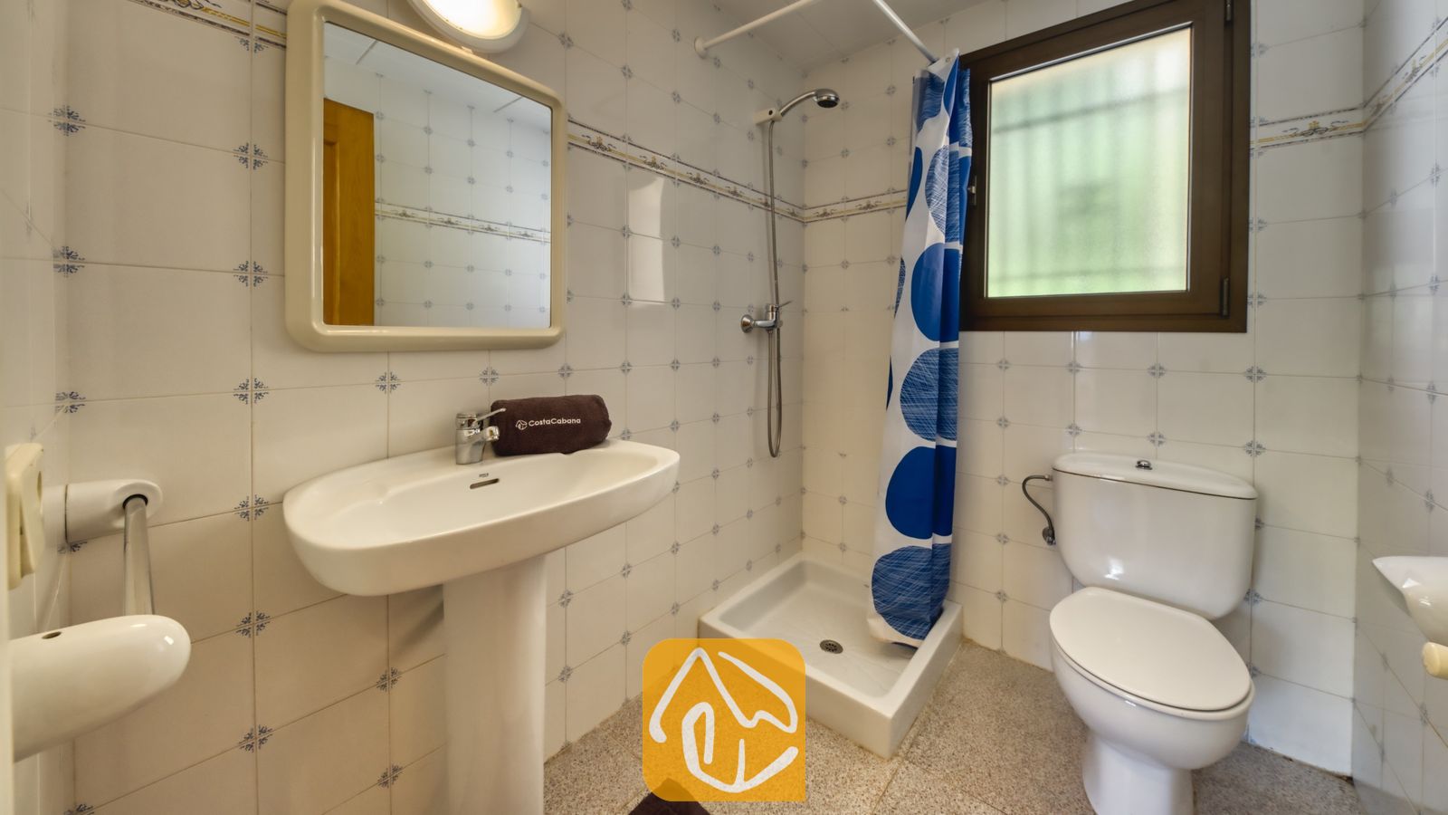 Vakantievilla's Costa Brava Spanje - Villa Paris - Wit betegelde badkamer met een wastafel op zuil, een wandspiegel, een hoekdouchecabine met een blauw gordijn met witte stippen, en een toilet.