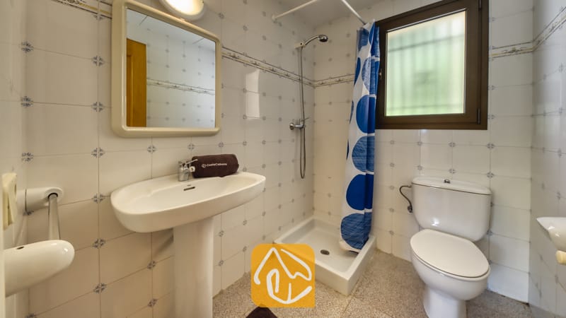 Villas de vacances Costa Brava Espagne - Villa Paris - Salle de bains carrelée de blanc comprenant un lavabo sur pied, un miroir mural, une cabine de douche d'angle avec un rideau à pois bleus et des toilettes.