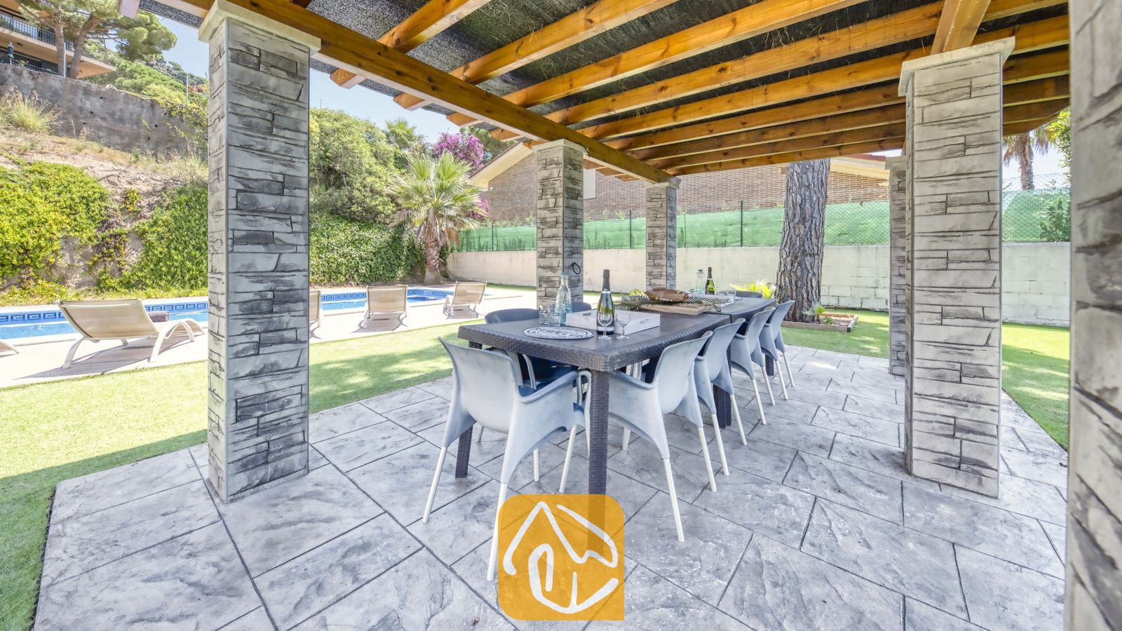 Vakantievilla's Costa Brava Spanje - Villa Paris - Overdekte eethoek buiten bij Villa Paris, voorzien van een donkere eettafel met kunststof stoelen onder een houten pergola gedragen door stenen pilaren.