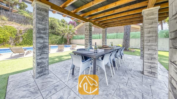 Vakantievilla's Costa Brava Spanje - Villa Paris - Overdekte eethoek buiten bij Villa Paris, voorzien van een donkere eettafel met kunststof stoelen onder een houten pergola gedragen door stenen pilaren.