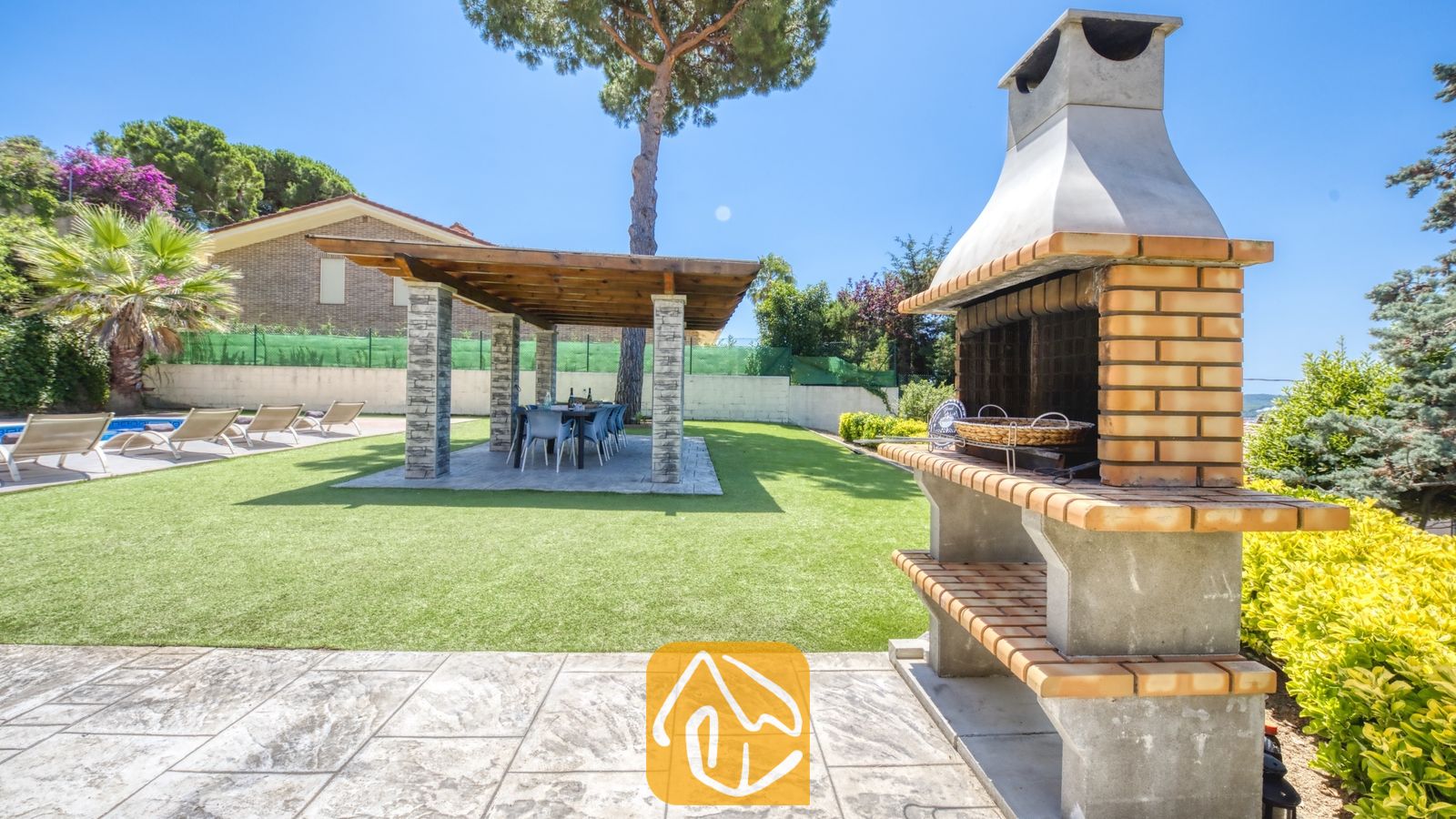 Vakantievilla's Costa Brava Spanje - Villa Paris - Stenen en betonnen buitenbarbecue op een stenen terras bij Villa Paris, met uitzicht op een groene grasmat met een overdekte pergola en eettafel.