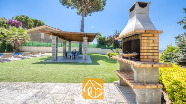 Vakantievilla's Costa Brava Spanje - Villa Paris - Stenen en betonnen buitenbarbecue op een stenen terras bij Villa Paris, met uitzicht op een groene grasmat met een overdekte pergola en eettafel.