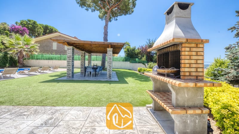 Vakantievilla's Costa Brava Spanje - Villa Paris - Stenen en betonnen buitenbarbecue op een stenen terras bij Villa Paris, met uitzicht op een groene grasmat met een overdekte pergola en eettafel.