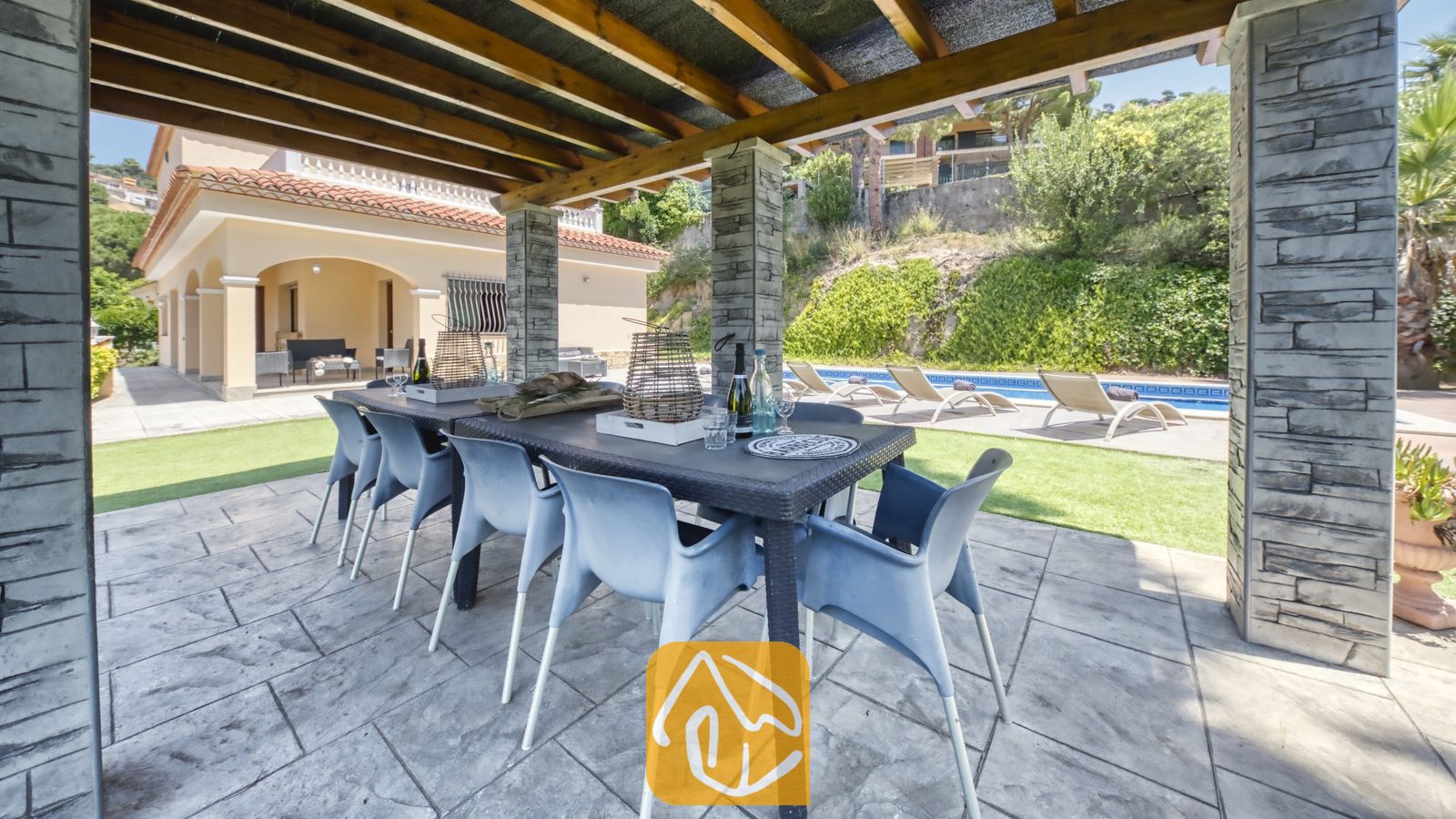Vakantievilla's Costa Brava Spanje - Villa Paris - Overdekte eethoek buiten bij Villa Paris, voorzien van een donkere kunststof tafel met grijskleurige stoelen, met natuursteen beklede zuilen en een houten pergola, met uitzicht op het zwembad en het terras.