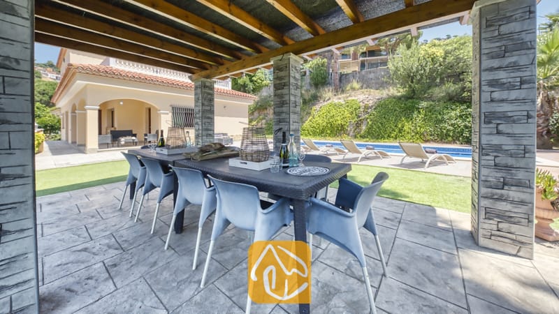 Vakantievilla's Costa Brava Spanje - Villa Paris - Overdekte eethoek buiten bij Villa Paris, voorzien van een donkere kunststof tafel met grijskleurige stoelen, met natuursteen beklede zuilen en een houten pergola, met uitzicht op het zwembad en het terras.