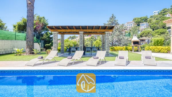 Vakantievilla's Costa Brava Spanje - Villa Paris - Het buitenzwembad van Villa Paris, voorzien van een rij witte ligbedden op het stenen terras, een overdekte eethoek onder een pergola en een ingebouwde buitenbarbecue op het gras.