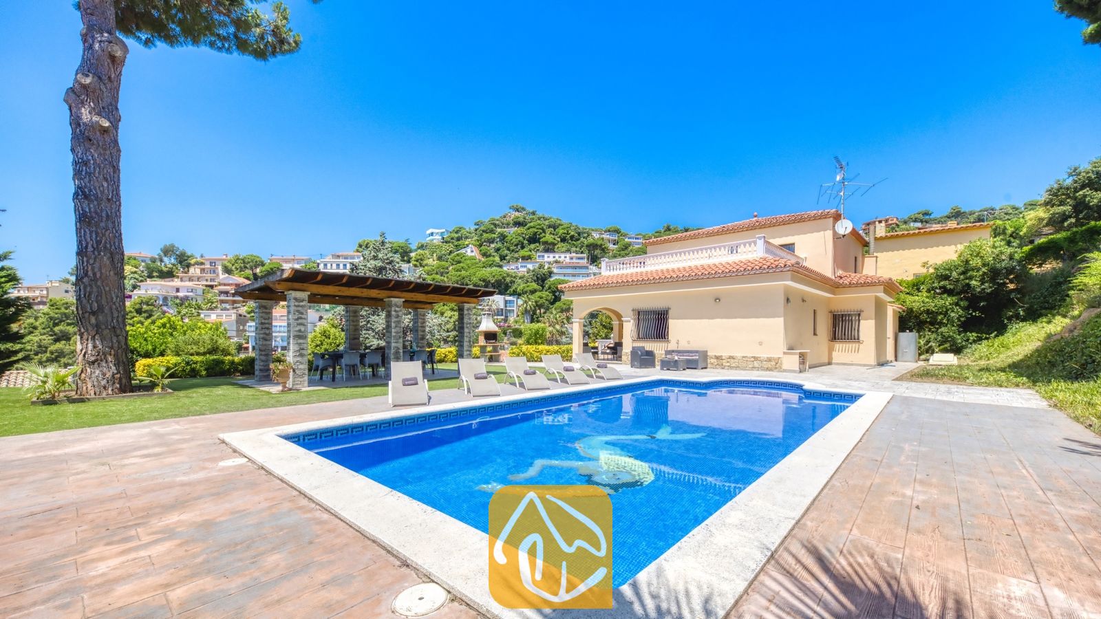Vakantievilla's Costa Brava Spanje - Villa Paris - Het buitenzwembad van Villa Paris met helderblauw water, ligstoelen op het stenen terras en een overdekte pergola langs het beige twee verdiepingen tellende huis.
