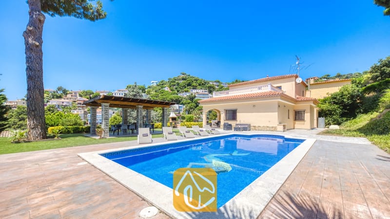 Vakantievilla's Costa Brava Spanje - Villa Paris - Het buitenzwembad van Villa Paris met helderblauw water, ligstoelen op het stenen terras en een overdekte pergola langs het beige twee verdiepingen tellende huis.