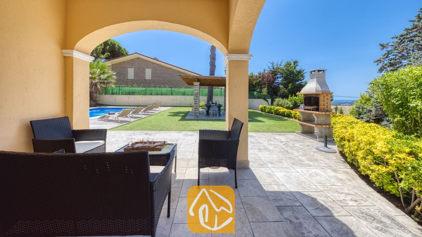 Vakantievilla's Costa Brava Spanje - Villa Paris - Overdekt terras bij Villa Paris met donkerwicker tuinmeubilair voorzien van witte kussens, uitkijkend over een betegeld terras, een groen gazon, een zwembad en een stenen barbecue.