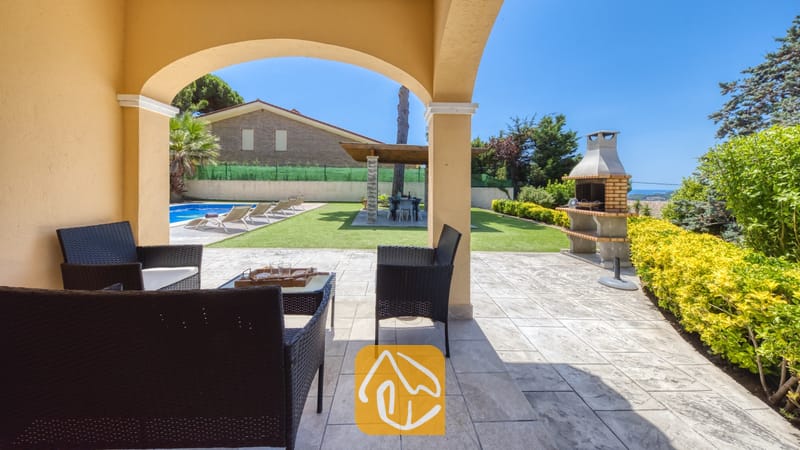 Vakantievilla's Costa Brava Spanje - Villa Paris - Overdekt terras bij Villa Paris met donkerwicker tuinmeubilair voorzien van witte kussens, uitkijkend over een betegeld terras, een groen gazon, een zwembad en een stenen barbecue.