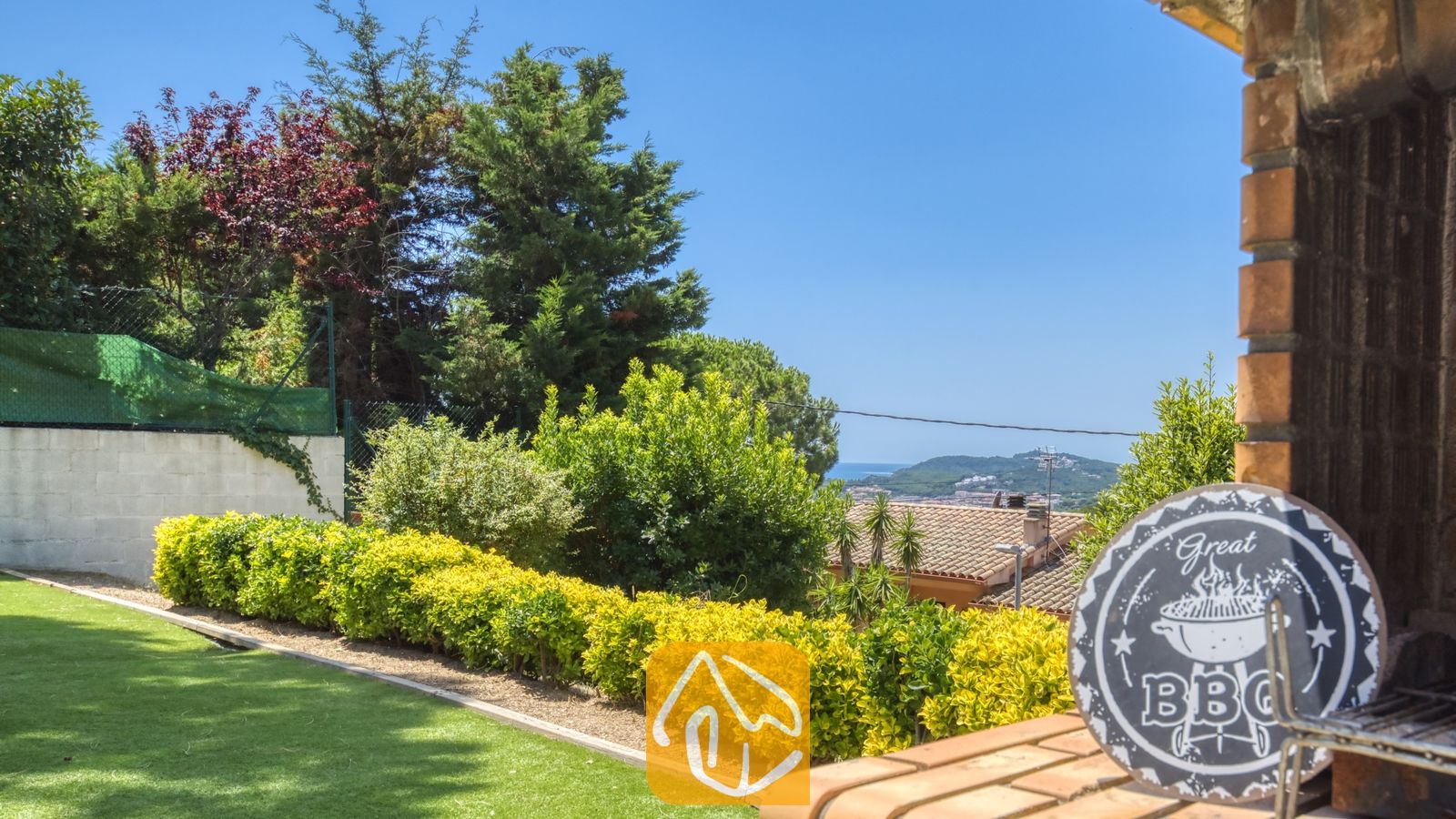 Vakantievilla's Costa Brava Spanje - Villa Paris - Buitengedeelte met stenen barbecue bij Villa Paris, voorzien van een decoratief BBQ-bord, verzorgde groene hagen en een uitzicht op de verre kustlijn onder een heldere, blauwe hemel.