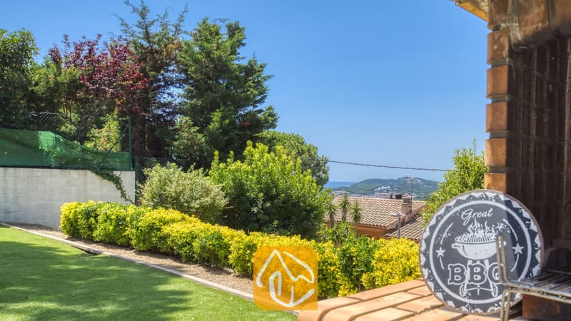 Vakantievilla's Costa Brava Spanje - Villa Paris - Buitengedeelte met stenen barbecue bij Villa Paris, voorzien van een decoratief BBQ-bord, verzorgde groene hagen en een uitzicht op de verre kustlijn onder een heldere, blauwe hemel.