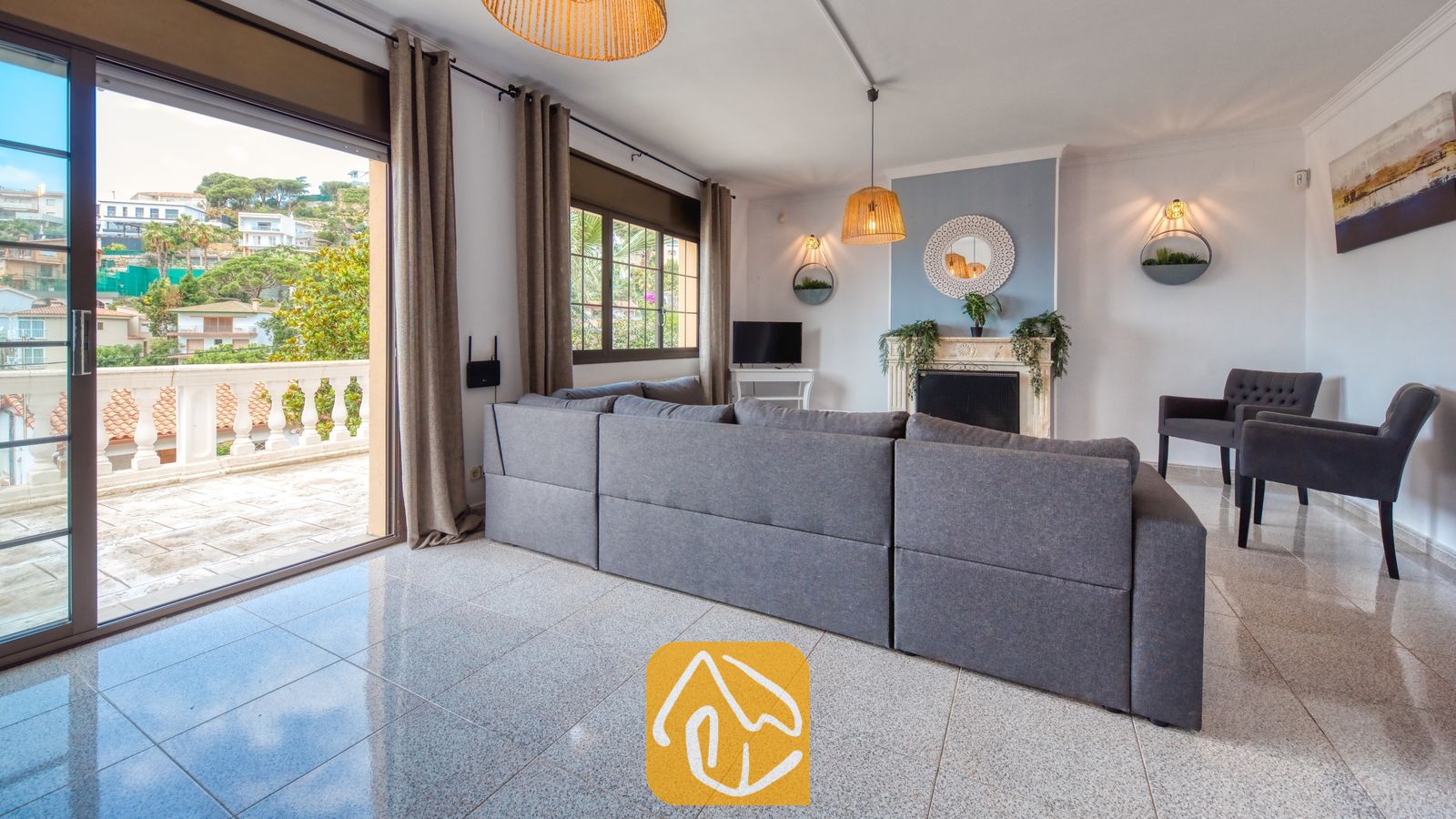 Vakantievilla's Costa Brava Spanje - Villa Paris - Ruime woonkamer in Villa Paris, voorzien van een grote grijze hoekbank, tegelvloeren en glazen schuifpuien die uitkomen op een terras met uitzicht op de heuvels.