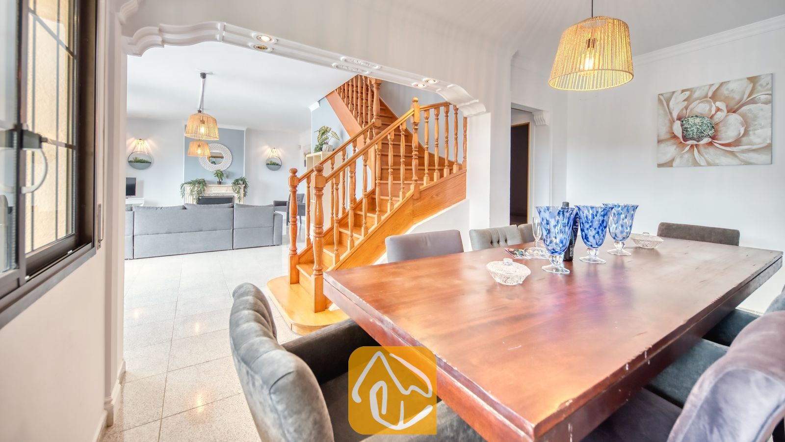 Vakantievilla's Costa Brava Spanje - Villa Paris - Houten eettafel met blauwe glazen op de voorgrond, met uitzicht op de woonkamer en de houten trap in Villa Paris