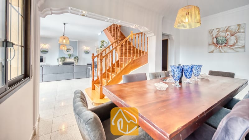 Villas de vacances Costa Brava Espagne - Villa Paris - Table à manger en bois avec des verres bleus au premier plan, donnant sur le salon et l'escalier en bois de la Villa Paris