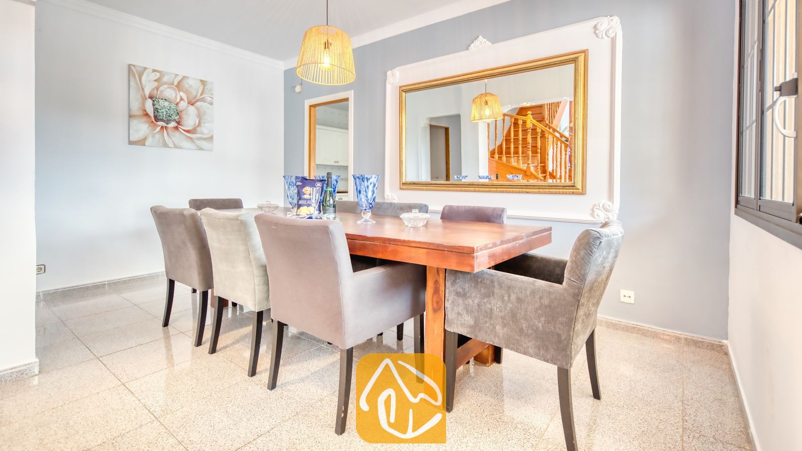 Vakantievilla's Costa Brava Spanje - Villa Paris - Eetkamer met een houten tafel omringd door grijze gestoffeerde fauteuils, een grote, sierlijke spiegel met gouden lijst en een hangende, geweven hanglamp.