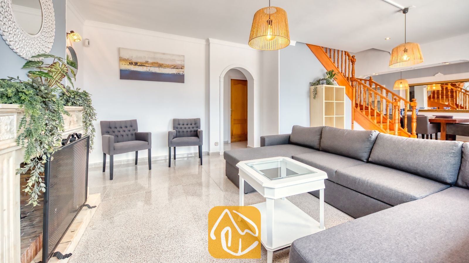 Vakantievilla's Costa Brava Spanje - Villa Paris - Interieur van de woonkamer in Villa Paris met een grijze hoekbank, een witte salontafel, geweven hanglampen en een open haard met groenvoorziening.