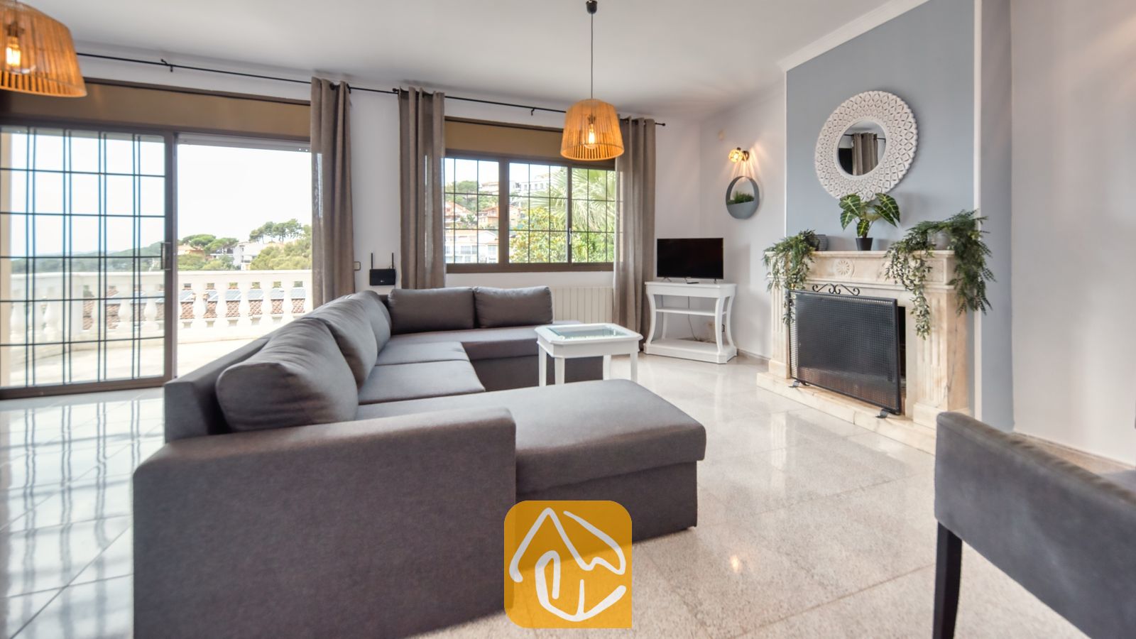 Vakantievilla's Costa Brava Spanje - Villa Paris - Woonkamer van Villa Paris met een grote grijze hoekbank, een witte open haard met decoratieve spiegel, tegelvloer en schuifpui naar een terras.