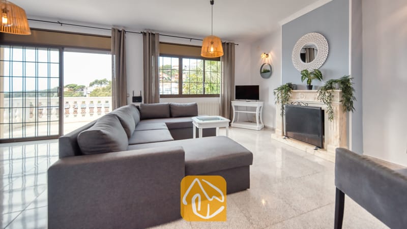 Vakantievilla's Costa Brava Spanje - Villa Paris - Woonkamer van Villa Paris met een grote grijze hoekbank, een witte open haard met decoratieve spiegel, tegelvloer en schuifpui naar een terras.