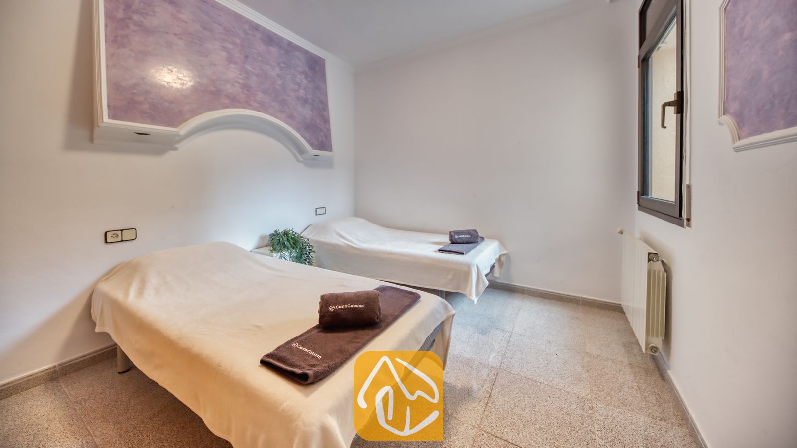 Vakantievilla's Costa Brava Spanje - Villa Paris - Slaapkamer met twee eenpersoonsbedden opgemaakt met wit linnengoed, opgevouwen bruine handdoeken, tegelvloeren en een paarse accentwand.
