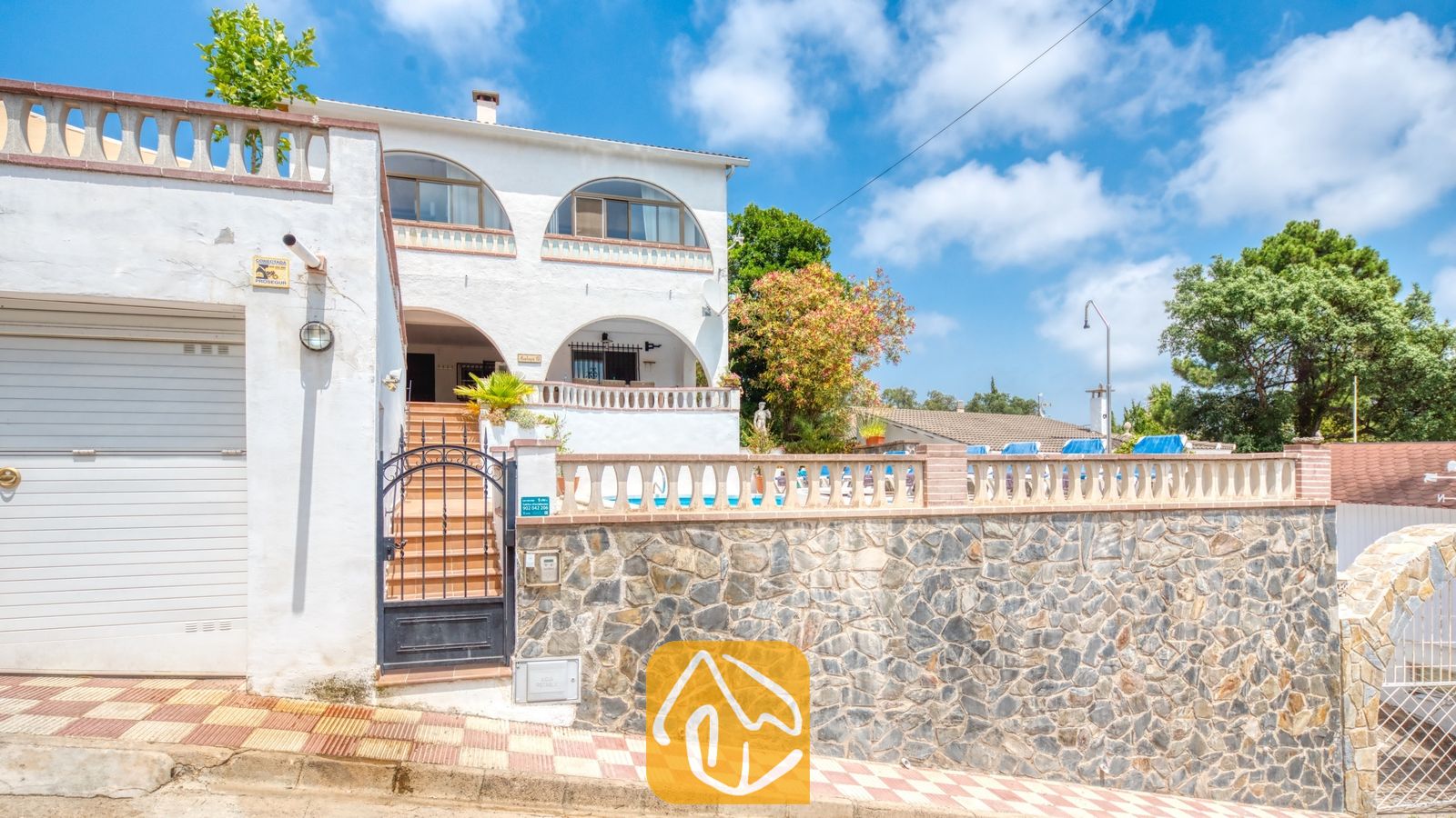 Vakantievilla's Costa Brava Spanje - Villa Santa Maria - Buitenaanzicht van Villa Santa Maria met een witte stucwerk gevel, een stenen muur, boogvensters en een afgesloten ingang die leidt naar een bovenste terras onder een blauwe hemel.
