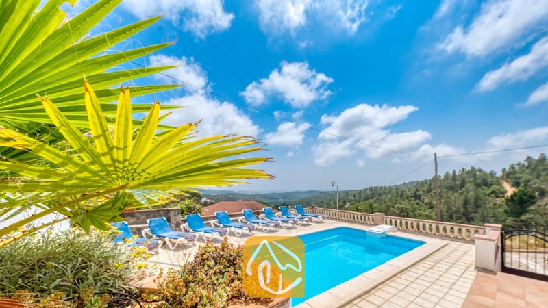 Vakantievilla's Costa Brava Spanje - Villa Santa Maria - Privézwembad met blauwe ligbedden en een tak van een palmprom op de voorgrond bij Villa Santa Maria onder een blauwe lucht met her en der wat wolken.
