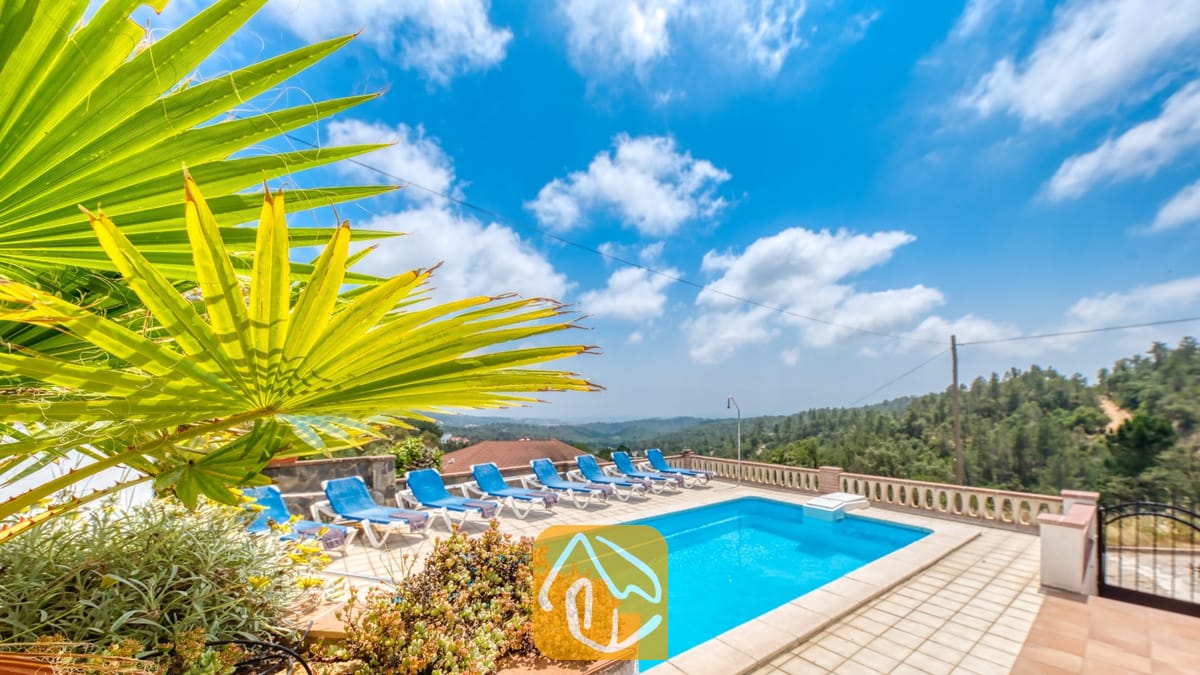 Vakantievilla's Costa Brava Spanje - Villa Santa Maria - Privézwembad met blauwe ligbedden en een tak van een palmprom op de voorgrond bij Villa Santa Maria onder een blauwe lucht met her en der wat wolken.