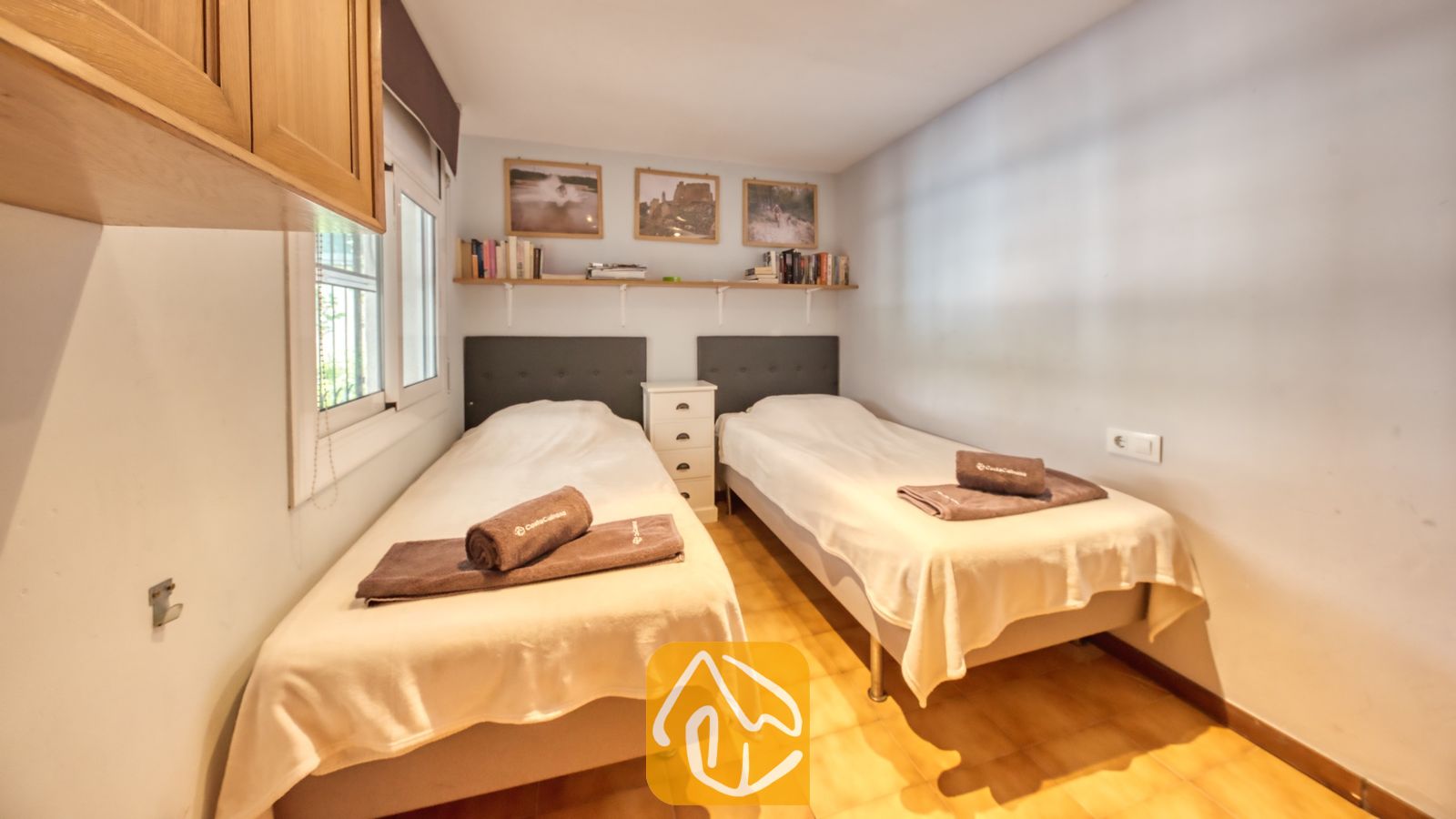 Vakantievilla's Costa Brava Spanje - Villa Santa Maria - Slaapkamer met twee eenpersoonsbedden voorzien van witte dekbedden, bruine opgevouwen handdoeken, een gedeeld wit nachtkastje, wandplanken met boeken en ingelijste foto's, en tegelvloeren.