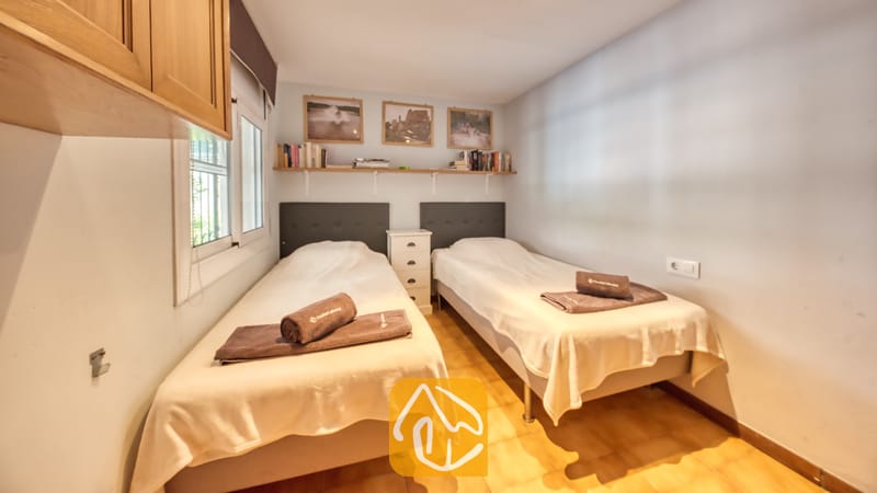 Villas de vacances Costa Brava Espagne - Villa Santa Maria - Chambre à deux lits simples dotée de couettes blanches, de serviettes pliées marron, d'une table de chevet blanche partagée, d'étagères murales avec des livres et des photos encadrées, et de sols carrelés.