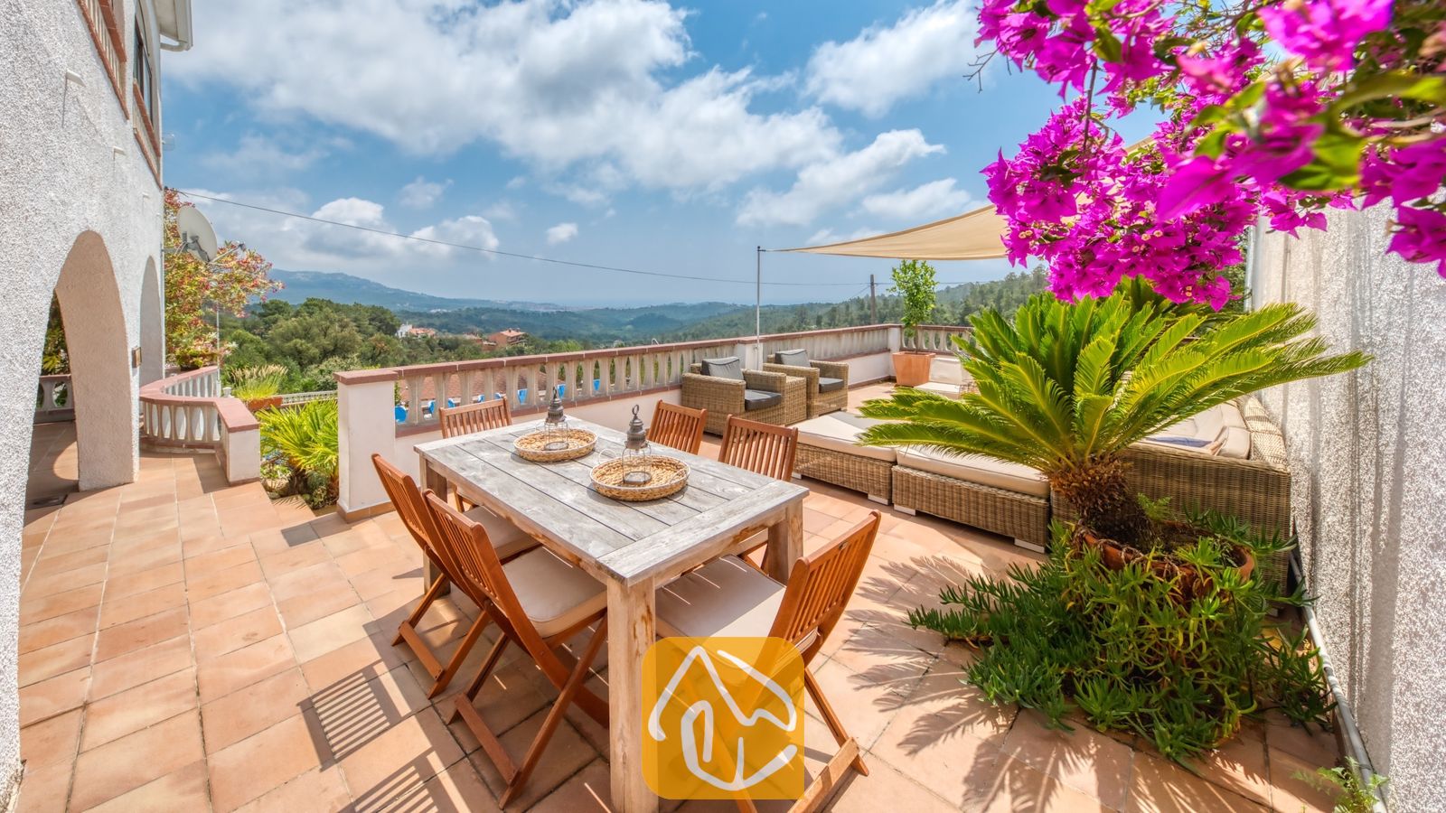 Vakantievilla's Costa Brava Spanje - Villa Santa Maria - Zonnig terras bij Villa Santa Maria, voorzien van een houten eettafel, rieten zitmeubelen en levendige roze bougainville met uitzicht op de verre heuvels.