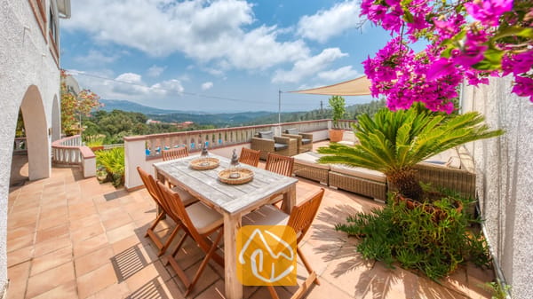 Vakantievilla's Costa Brava Spanje - Villa Santa Maria - Zonnig terras bij Villa Santa Maria, voorzien van een houten eettafel, rieten zitmeubelen en levendige roze bougainville met uitzicht op de verre heuvels.