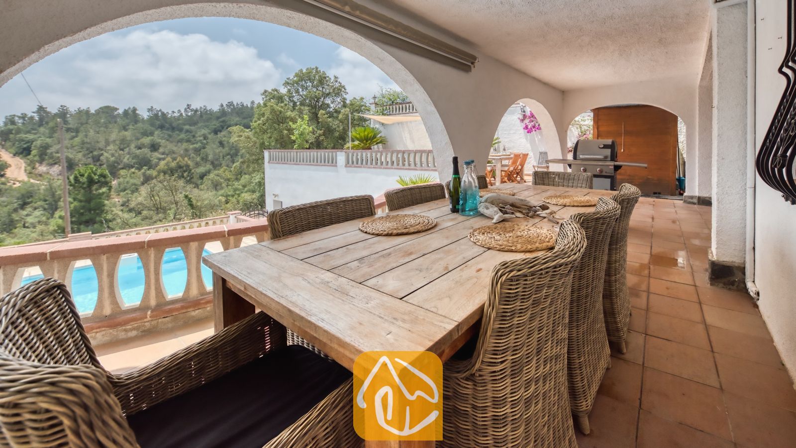 Vakantievilla's Costa Brava Spanje - Villa Santa Maria - Overdekt terras bij Villa Santa Maria, voorzien van een lange houten eettafel, rieten fauteuils en bogen die uitzicht bieden op de bomen en een zwembad.
