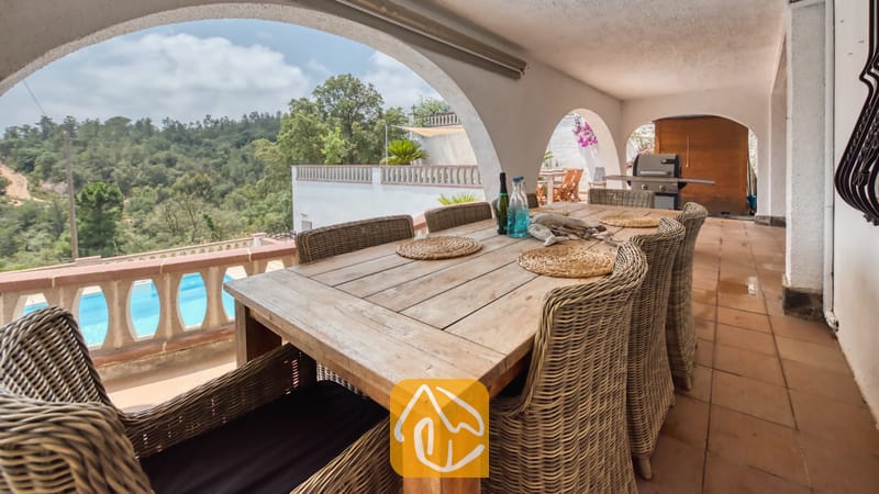 Vakantievilla's Costa Brava Spanje - Villa Santa Maria - Overdekt terras bij Villa Santa Maria, voorzien van een lange houten eettafel, rieten fauteuils en bogen die uitzicht bieden op de bomen en een zwembad.