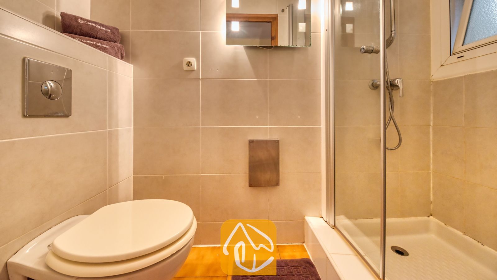 Vakantievilla's Costa Brava Spanje - Villa Santa Maria - Moderne badkamer met beige keramische wandtegels, een wit toilet en een glazen inloopdouche met chromen armatuur.
