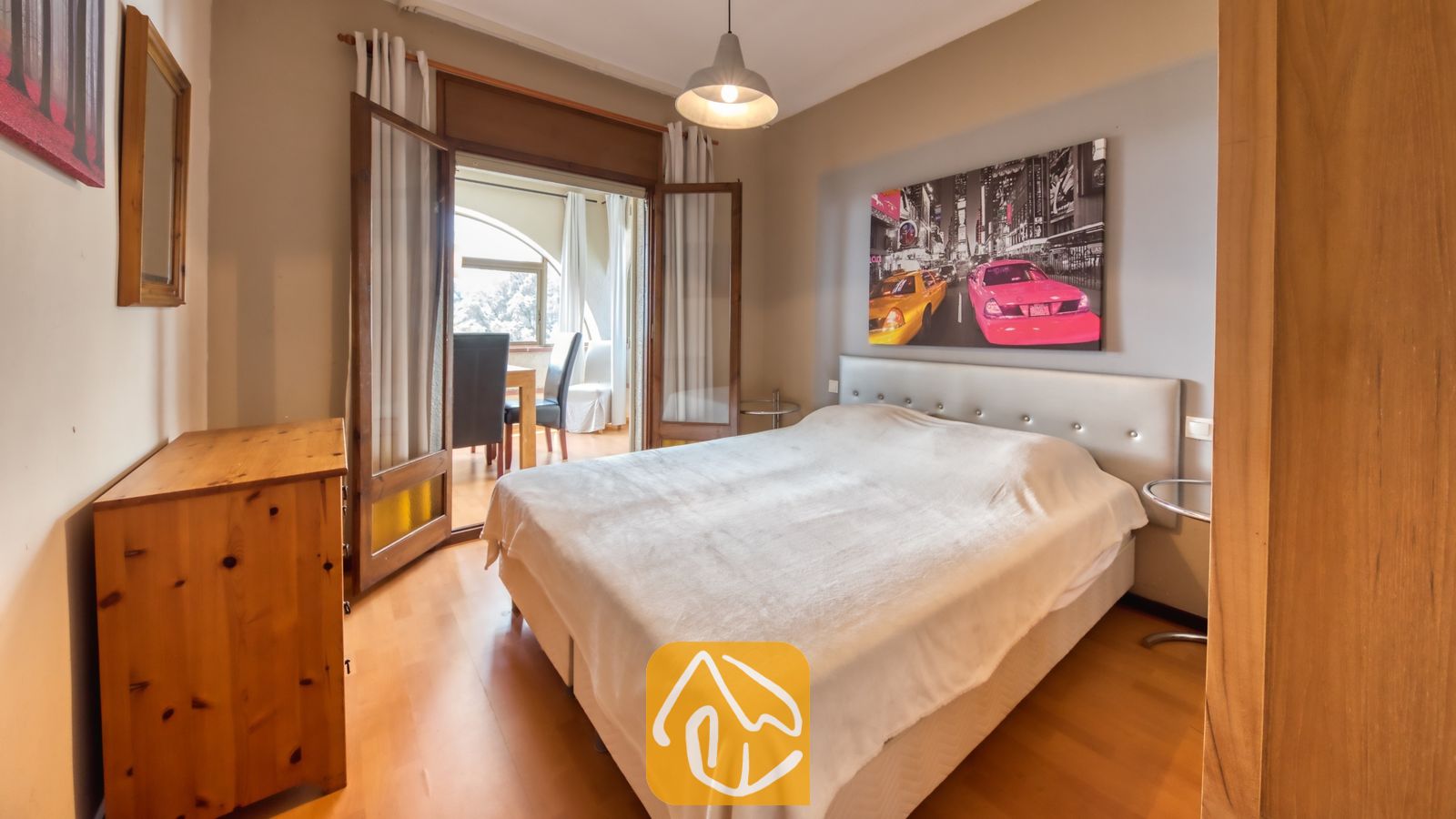 Vakantievilla's Costa Brava Spanje - Villa Santa Maria - Slaapkamer met een tweepersoonsbed, houten dressoir, hanglamp en open glazen deuren die naar een balkon leiden.