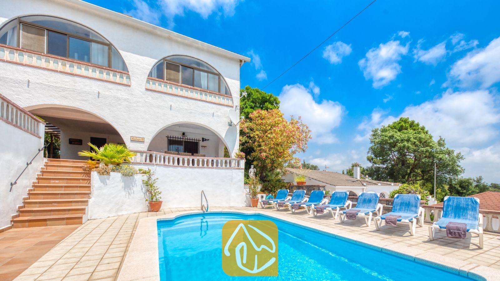 Vakantievilla's Costa Brava Spanje - Villa Santa Maria - Buitenzwembad en witte buitenkant van Villa Santa Maria in Spanje, met blauwe ligstoelen, boogvormige ramen en tegelいません en betegelde trappen onder een blauwe lucht met wolken.